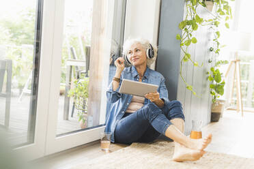 Reife Frau, die wegschaut und Musik hört, während sie an einem digitalen Tablet arbeitet - UUF21660