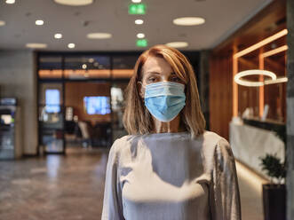 Senior woman wearing protective face mask standing in hotel lobby - ZEDF03830