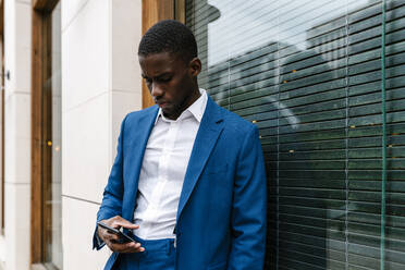 Junger männlicher Unternehmer, der sein Smartphone benutzt, während er sich in der Stadt ans Fenster lehnt - EGAF00798