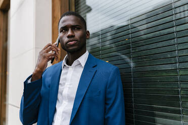Businessman looking away while listening through mobile phone during call leaning on window - EGAF00799