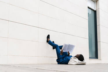 Männlicher Unternehmer liegt mit Seesack auf dem Gehweg und liest ein Buch in der Stadt - EGAF00801