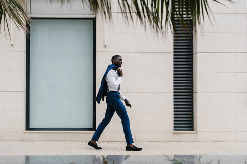 Lächelnder männlicher afrikanischer Berufstätiger, der in der Stadt auf dem Bürgersteig geht - EGAF00806