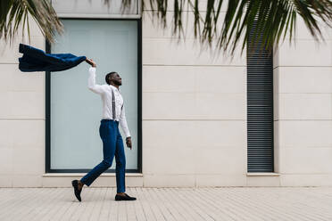 Happy male entrepreneur holding blue suit while walking on sidewalk in city - EGAF00807