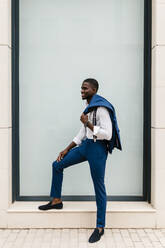 Lächelnder junger männlicher Unternehmer, der einen Anzug hält und am Fenster in der Stadt steht - EGAF00813