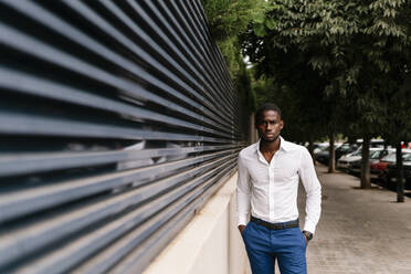Selbstbewusster afrikanischer Fachmann, der mit den Händen in den Taschen an einer Mauer in der Stadt steht - EGAF00838