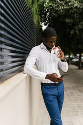 Geschäftsmann, der aus einer Tasse trinkt, während er sein Smartphone benutzt und sich an eine Wand lehnt - EGAF00840