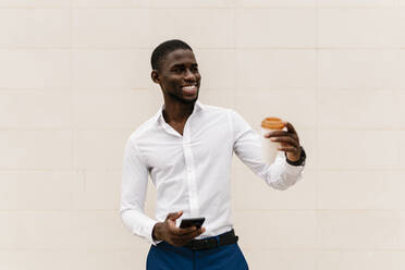 Lächelnder junger Geschäftsmann, der wegschaut, während er einen wiederverwendbaren Becher und ein Smartphone an eine Wand in der Stadt hält - EGAF00843