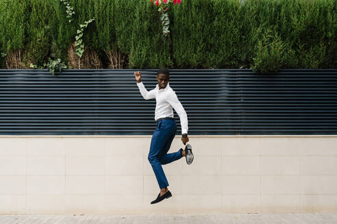 Male professional jumping on sidewalk against surrounding wall in city - EGAF00844