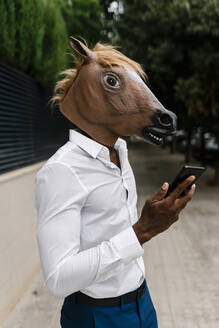 Male entrepreneur wearing horse mask while holding smart phone in city - EGAF00848