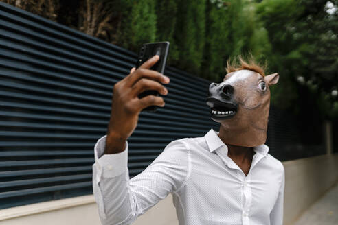Businessman wearing horse mask while taking selfie from smart phone in city - EGAF00849