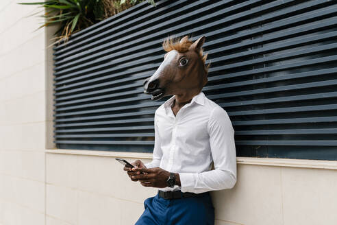 Young male African professional wearing horse mask while using smart phone against surrounding wall in city - EGAF00850