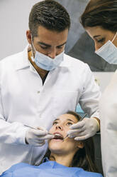 Male dentist in mask and gloves examining teeth of pretty patient with help of female assistant at clinic - ABZF03354