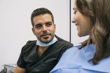 Gut aussehender männlicher Zahnarzt erklärt einer lächelnden Patientin in der Praxis die Zahnbehandlung - ABZF03364