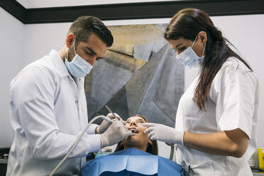 Male dentist in surgical glove and mask doing dental treatment of patient with help of female assistant - ABZF03373