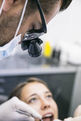Männlicher Zahnarzt mit chirurgischer Lupenbrille untersucht die Zähne einer Patientin in der Klinik - ABZF03377