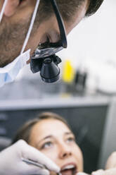 Male dentist in surgical loupes examining teeth of female patient in clinic - ABZF03377