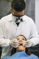 Männlicher Zahnarzt mit chirurgischer Lupenbrille bei der Untersuchung der Zähne einer jungen Patientin in der Klinik - ABZF03382