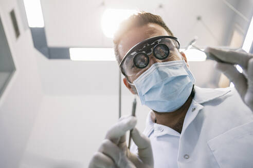 Männlicher Zahnarzt in chirurgischer Lupenbrille und Maske mit medizinischen Instrumenten während der Behandlung in der Klinik - ABZF03398