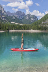 Man doing headstand on paddleboard at Pragser Wildsee against mountain range, Dolomites, Alto Adige, Italy - MMAF01419