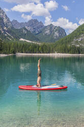 Mann macht Kopfstand auf dem Paddleboard am Pragser Wildsee vor der Bergkette, Dolomiten, Südtirol, Italien - MMAF01419