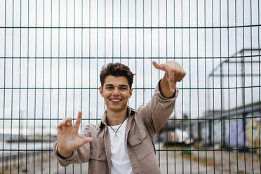 Lächelnder junger Mann mit Handzeichen am Metallzaun - DCRF00887