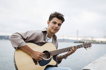 Hübscher junger Mann spielt Gitarre beim Üben an der Promenade gegen den Himmel - DCRF00890