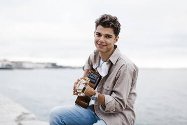 Lächelnder Mann spielt Gitarre beim Üben an der Promenade gegen den Himmel - DCRF00891