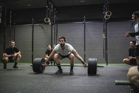 Athlete cheering man crouching while lifting barbell at gym stock photo