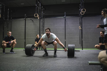 Sportler, der einen Mann anfeuert, der beim Heben einer Langhantel im Fitnessstudio hockt - SNF00559