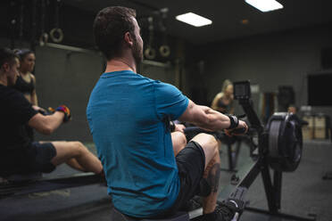 Mann benutzt Rudergerät mit Menschen, die im Hintergrund im Fitnessstudio trainieren - SNF00567