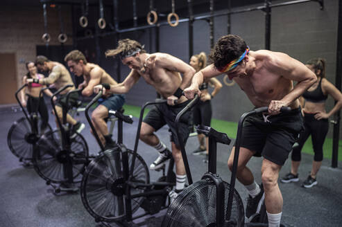 Athletes cycling on exercise bike with women standing in background at gym - SNF00571