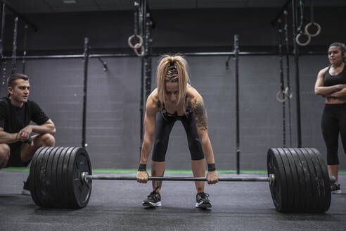 Athleten beobachten Frau, die im Fitnessstudio stehend eine Langhantel nimmt - SNF00584