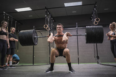 Athletes cheering man crouching while picking barbell up at gym - SNF00587