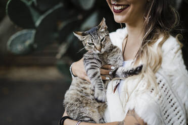 Lächelnde Frau, die eine Katze hält, während sie gegen eine Pflanze steht - DCRF00919