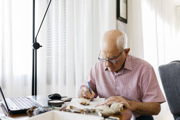 Älterer Mann schreibt in ein Buch und sitzt mit Mineralien und Fossilien am Tisch - JRFF04757