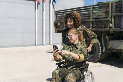 Lächelnde behinderte Soldatin, die ein Selfie mit einem jungen Kollegen vor einem Lastwagen auf einem Armeestützpunkt an einem sonnigen Tag macht - MTBF00668