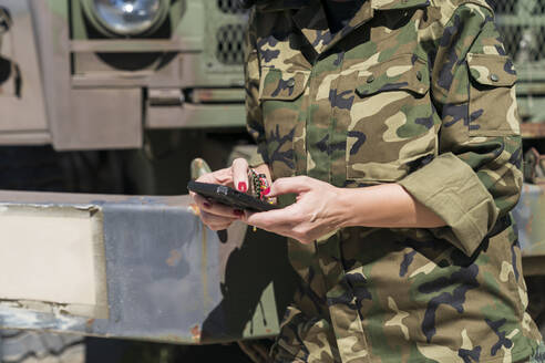 Älterer weiblicher Armeesoldat, der ein Mobiltelefon benutzt, während er an einem sonnigen Tag gegen einen Lastwagen in einem Stützpunkt steht - MTBF00675