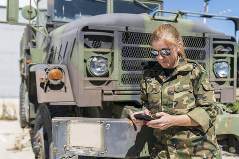 Mature blond female army soldier text messaging through smart phone while standing against truck at base on sunny day - MTBF00676