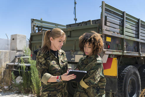 Multi-ethnic female army soldiers discussing over digital tablet at military base on sunny day - MTBF00677