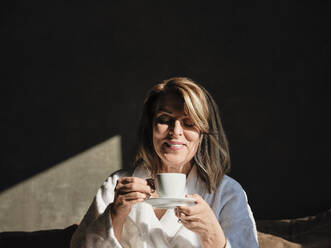 Smiling blond woman enjoying fresh coffee while sitting in hotel room - ZEDF03869