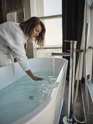 Ältere Frau beugt sich vor und prüft die Temperatur in der Badewanne eines Luxushotels - ZEDF03874