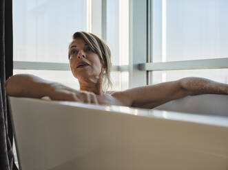 Blonde ältere Frau nimmt ein Bad in der Badewanne vor dem Fenster eines Luxushotels - ZEDF03879