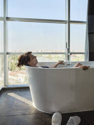 Entspannte ältere Frau im Ruhestand, die ein Bad in einer Badewanne vor dem Fenster eines Luxushotels nimmt - ZEDF03885