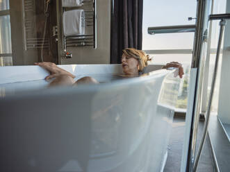 Retired woman sleeping while taking bath in bathtub against window at luxury hotel room - ZEDF03888