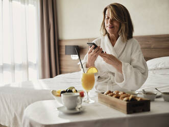 Lächelnde blonde ältere Frau, die ihr Smartphone benutzt, während sie beim Frühstück im Hotelzimmer sitzt - ZEDF03889
