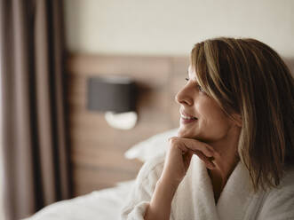 Thoughtful smiling blond woman looking away while sitting in hotel room - ZEDF03895