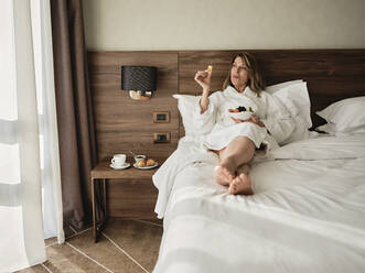 Senior woman eating fresh fruits in breakfast while relaxing on bed and looking away at hotel room - ZEDF03899