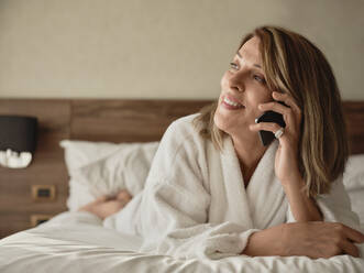 Lächelnde ältere Frau, die am Telefon spricht, während sie wegschaut und auf dem Bett im Hotelzimmer liegt - ZEDF03907