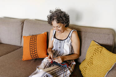 Lächelnde Frau beim Stricken auf dem Sofa zu Hause sitzend - TCEF01192