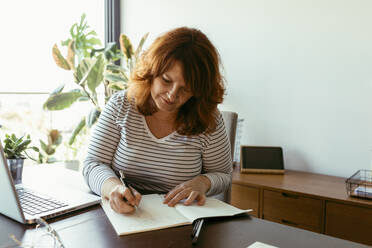 Frau, die in einem Boo schreibt, während sie einen Laptop auf einem Tisch zu Hause benutzt - VABF03557